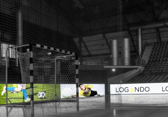 Leeres Hallenfußballtor auf einem glänzenden Hallenboden; im Hintergrund digitale LED-Banden mit Werbung, u.a. für das LED-Bandenmodul 10.00 mm - Memphiz 10 mieten. Die Tribünen sind größtenteils ohne Zuschauer. Ideal für Interessierte an Fußballtor-Miete oder Arena-Flächenvermietung.