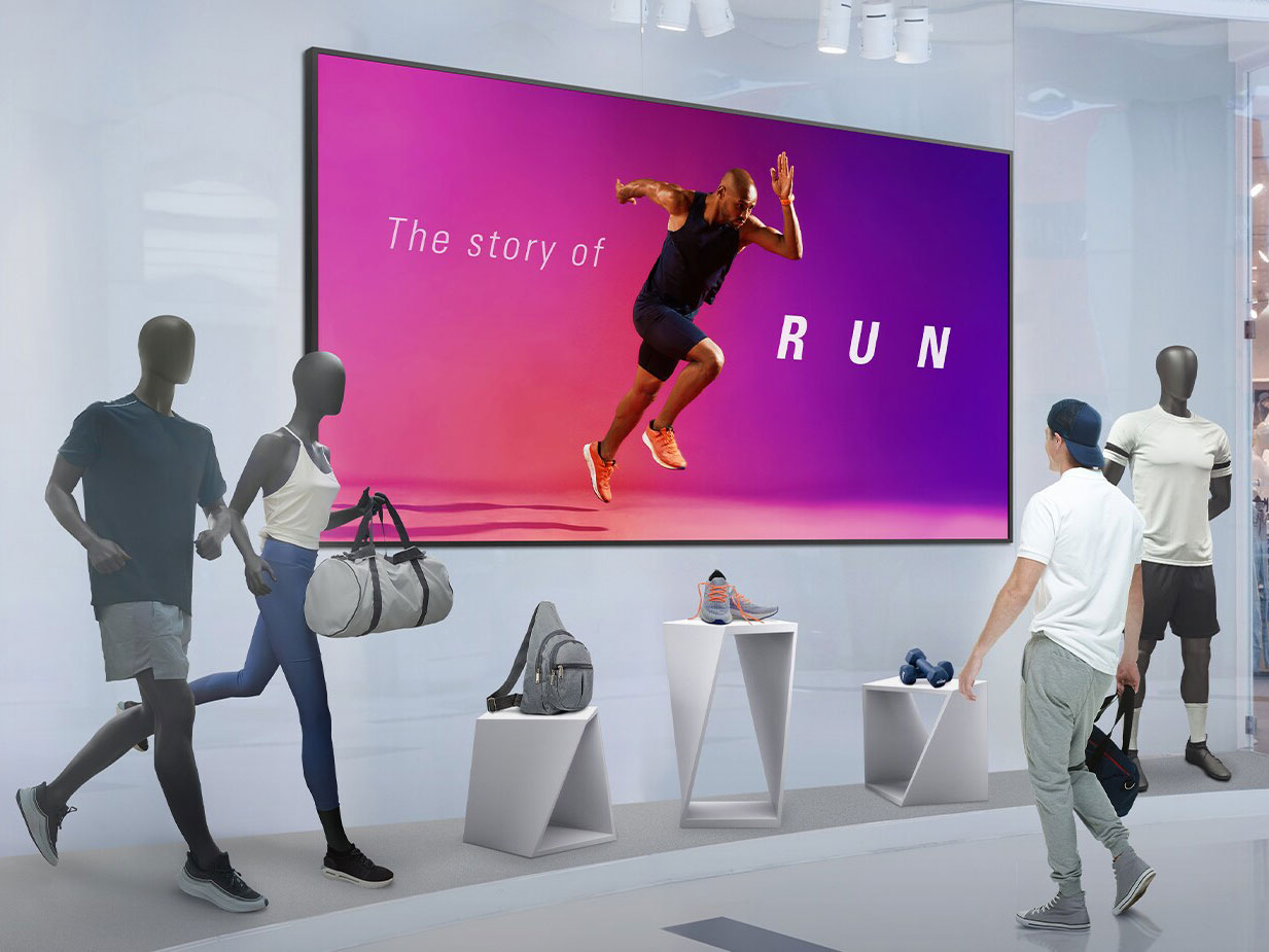 Ein Mann in Sportkleidung steht in einem modernen Geschäft vor einem 110 Zoll Digital Signage Display - LG 110UM5J-B, das ein Bild eines Läufers und den Schriftzug "The story of RUN" zeigt. Im Hintergrund präsentieren Schaufensterpuppen verschiedene Sportoutfits zur Miete oder zum Kauf.