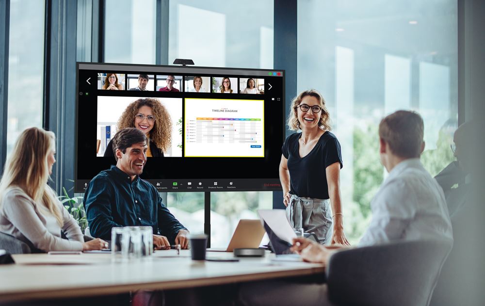 Vier Personen sitzen lächelnd an einem Konferenztisch und sprechen miteinander, während eine Frau auf einem 55 Zoll UHD Display Optoma N3551K (Neuware) Präsentationsoptionen zum Mieten oder Kaufen zeigt. Helle Fenster im Hintergrund spenden natürliches Licht während einer virtuellen Besprechung.