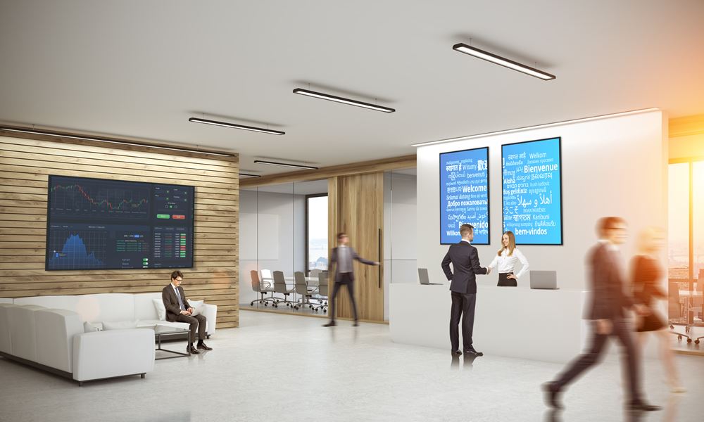 Modern eingerichtete Büro-Lobby mit Menschen, die am Empfangsschalter interagieren; ein Mann sitzt auf einem weißen Sofa. Im Vordergrund steht ein Optoma N3551K 55 Zoll UHD Display (Neuware), ideal für Unternehmen. Im Hintergrund ist ein Konferenzraum zu sehen; Sonnenlicht fällt durch große Fenster.