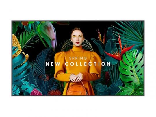 Eine Frau im orangefarbenen Kleid mit passender Tasche steht vor bunten tropischen Blättern und Blumen. Im Vordergrund ist auf dem Bildschirm der Text "85 Zoll Smart Signage Display - Samsung QB85C mieten - jetzt mieten oder kaufen" zu sehen. Das Produktbild zeigt das große Samsung QB85C Smart Signage Display, ideal für digitale Werbepräsentationen.