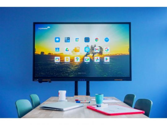 Das 55 Zoll Touchdisplay Legamaster EVOLVE 2 ETX-5540 (Neuware) zeigt farbige App-Icons vor einer blauen Wand in einem modernen Meetingraum. Vor dem Display steht ein Tisch mit Laptops, Notizbüchern, Stiften und Kaffeetassen sowie grünen Stühlen.