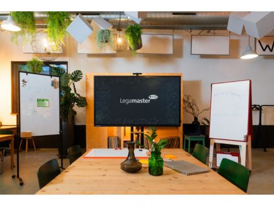 Das Produktbild zeigt ein modernes Konferenzzimmer mit einem 65 Zoll Touchdisplay Legamaster EXPLORE 2 EXP-6510 (Neuware) an der Wand, umgeben von Whiteboards und Pflanzen. Im Vordergrund steht ein Holztisch mit Vasen und Markern, grüne Stühle sind ringsum angeordnet. Die grüne Bestuhlung sowie die Präsentationstechnik können gekauft oder gemietet werden.