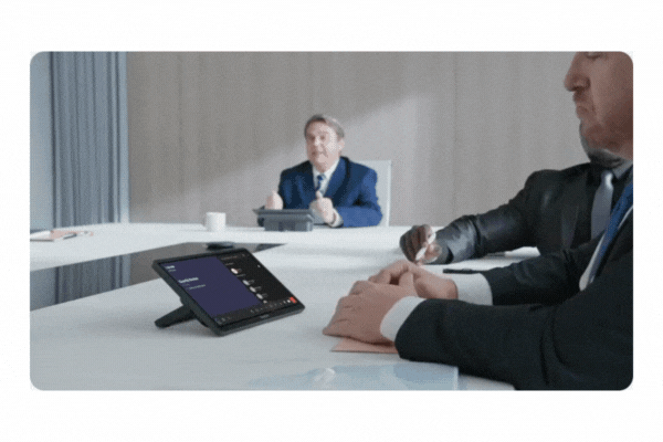 Geschäftsleute besprechen in einem modernen Konferenzraum den Kauf der 11.6" Touch Console - Yealink MTouch Plus (Neuware); im Vordergrund zeigt ein Tablet mit großem Touchscreen die Videokonferenz-Oberfläche, während ein Mann im blauen Anzug gestikulierend spricht.