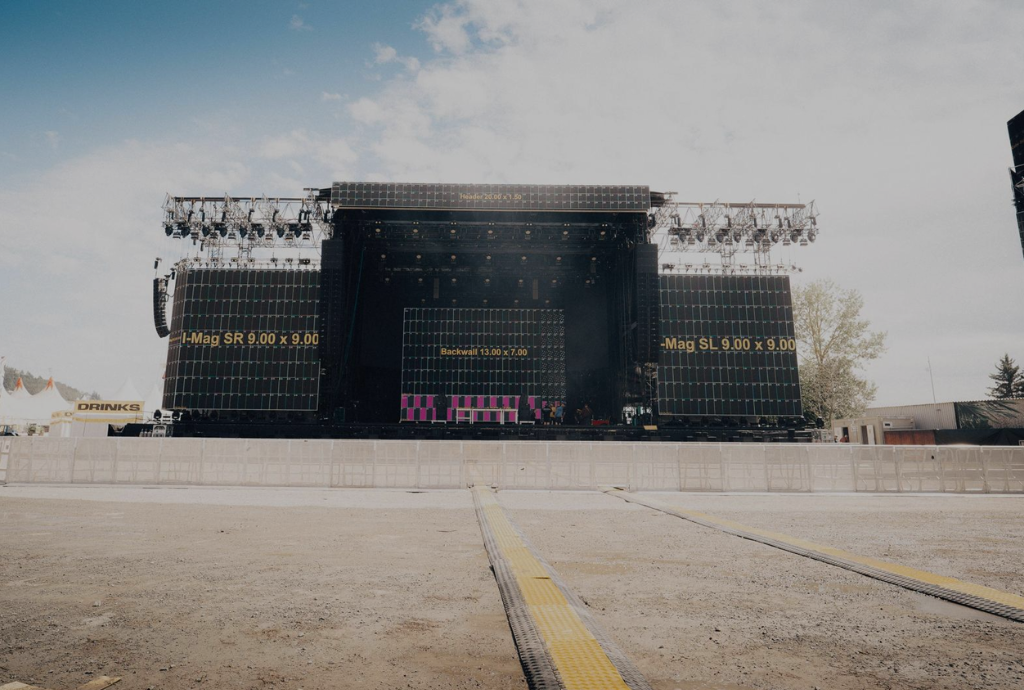 OpenAir Festivalbühne: Große Bühne mit LED-Leinwänden & Licht, leer, unter Wolkenhimmel, Bäume und Festival-Schilder im Hintergrund.